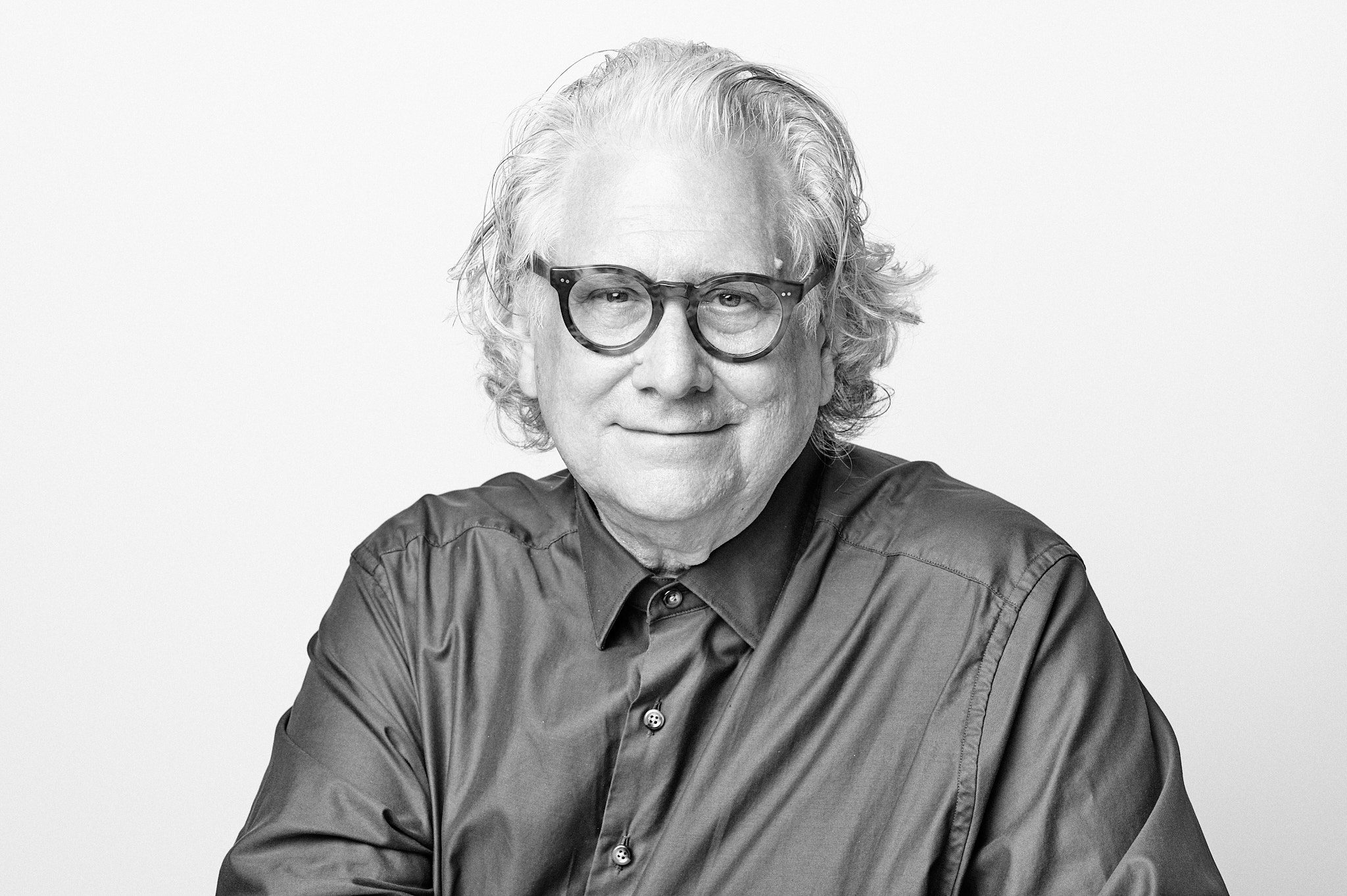 Black-and-white portrait of an older man with wavy hair and round glasses, wearing a button-down shirt. He smiles gently at the camera against a plain white background.