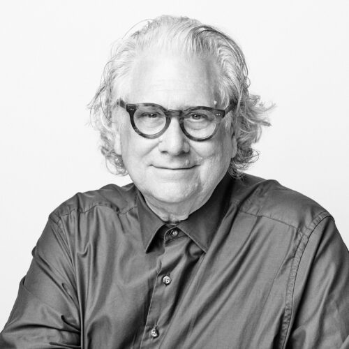 Black-and-white portrait of an older man with wavy hair and round glasses, wearing a button-down shirt. He smiles gently at the camera against a plain white background.