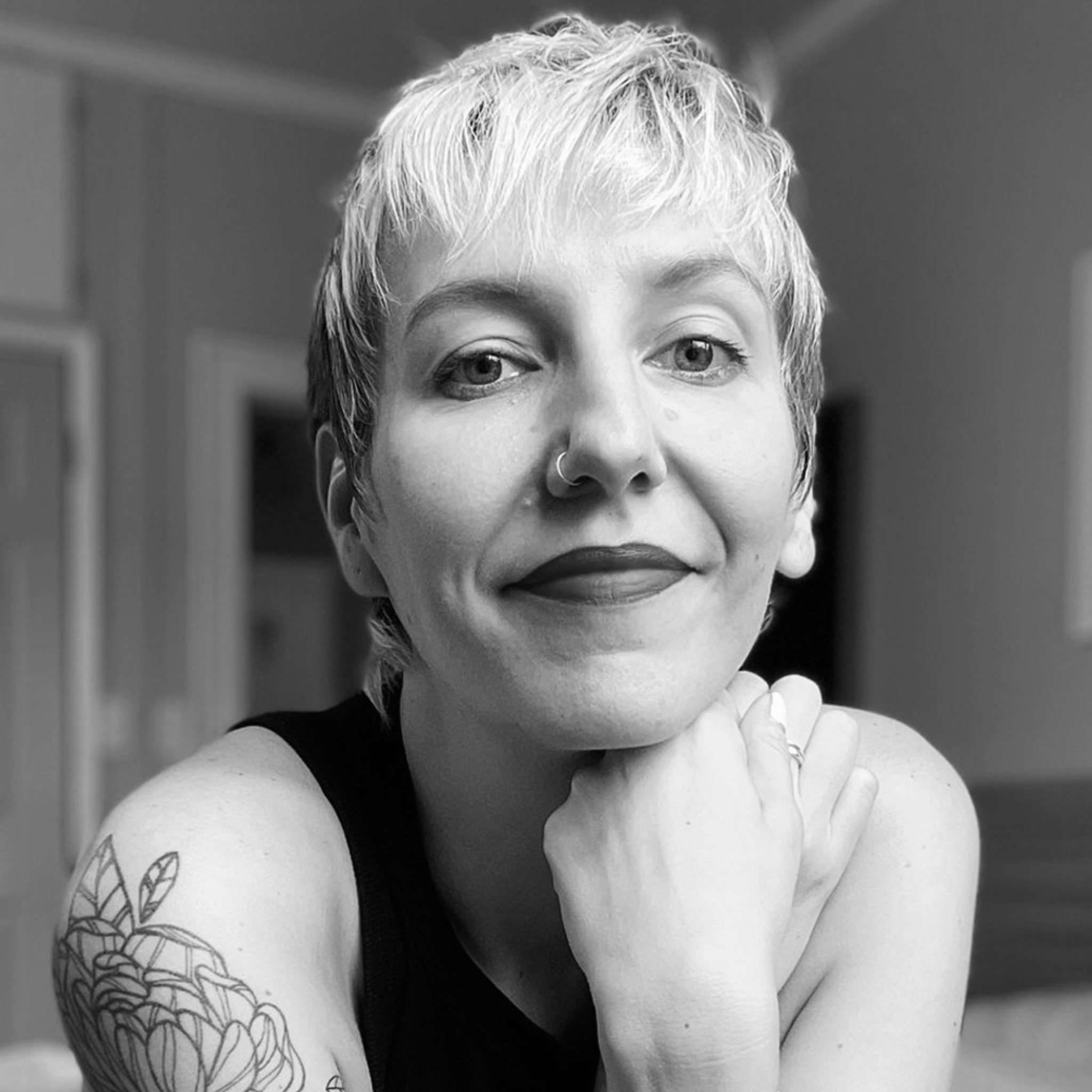 A woman with short, platinum blonde hair, a nose ring, and visible floral tattoos rests her chin on her hand. She wears a sleeveless top and smiles slightly in a relaxed, candid moment. The background is softly blurred.
