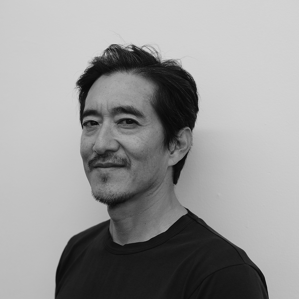 A man with short dark hair and a neatly trimmed goatee stands in front of a white wall. He wears a plain dark shirt and gazes toward the camera with a relaxed, confident expression. The image is in black and white.