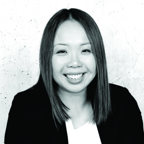 A grayscale image of a woman with shoulder-length curly hair, smiling brightly. She wears a soft sweater, and the photo is warmly lit, with the background softly blurred for focus on the subject’s friendly expression.