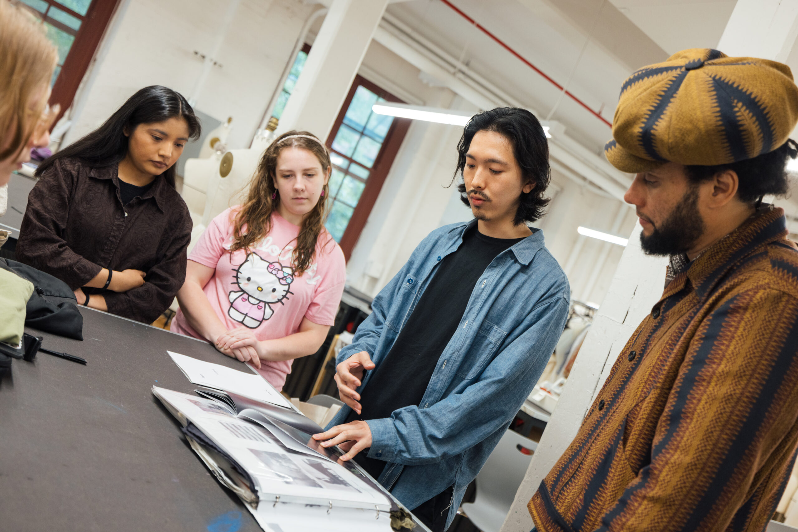 A group of people stand around a table in a fashion studio while one person gestures to an open book, discussing design work in a collaborative classroom setting.