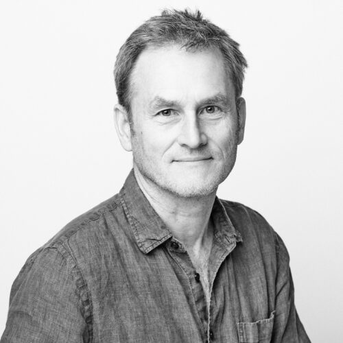 Black-and-white portrait of a man with short hair and a slight beard, wearing a casual button-down shirt. He smiles softly at the camera against a plain white background.