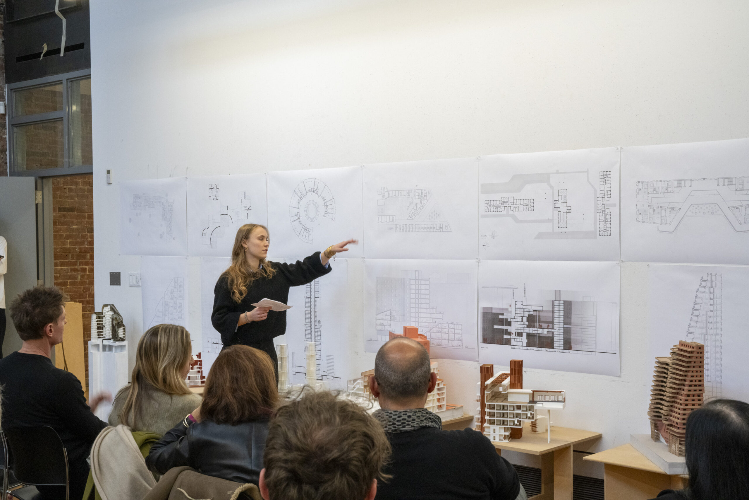 A student stands at the front of a room presenting architectural drawings mounted on a wall. She gestures toward the plans while holding papers in her other hand. Several audience members sit facing her, and scale architectural models are displayed on tables in the foreground.