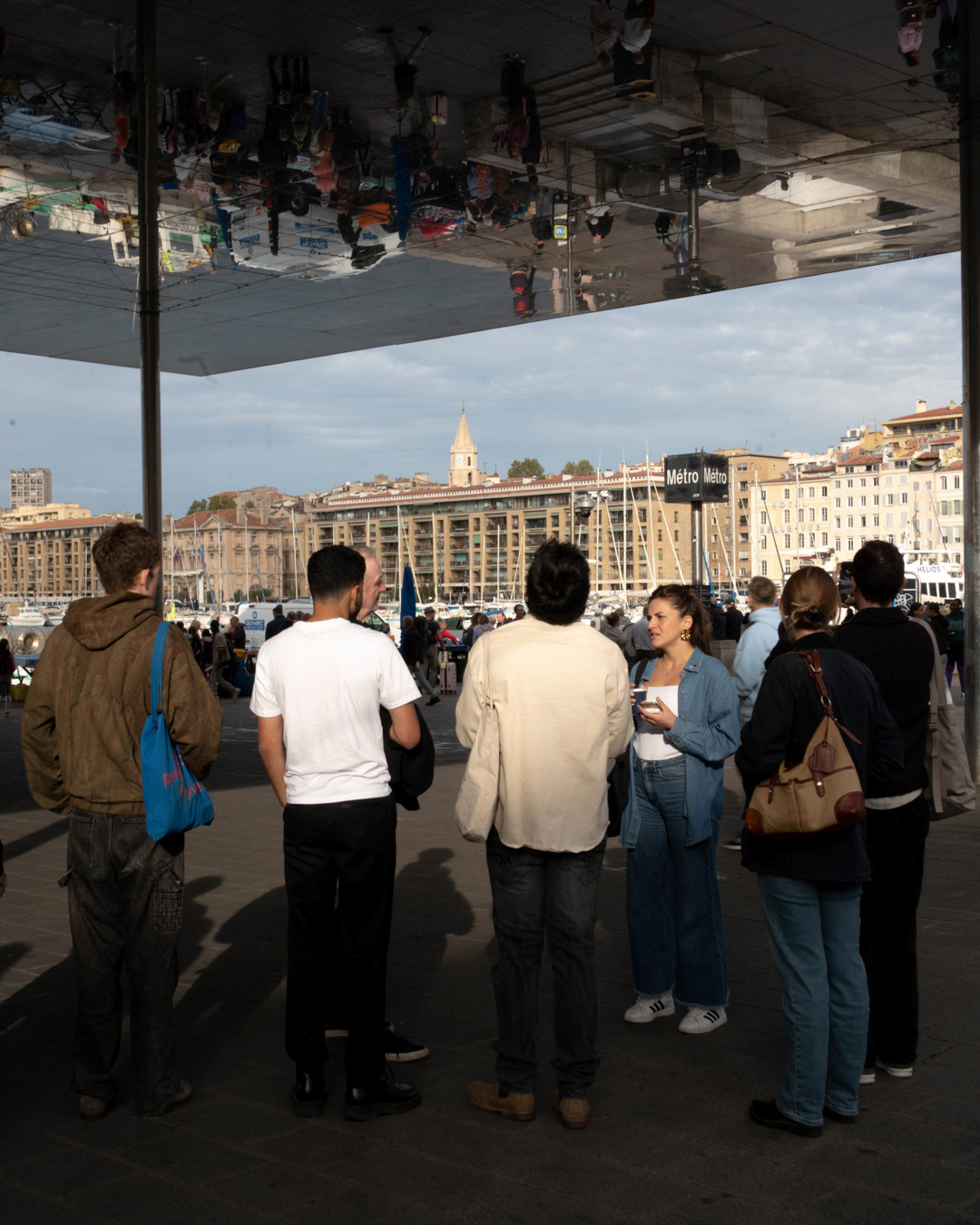 A group of people stand outdoors near a waterfront under a large reflective canopy. The canopy mirrors the scene above them. In the background, buildings, boats, and a sign reading “Métro” are visible. The group appears to be engaged in conversation while facing the harbor.