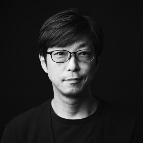 A man with short hair and glasses looks directly into the camera with a calm expression. He wears a dark crew neck shirt. The background is pitch black, drawing focus to the subject’s face in this sharply lit black and white portrait.