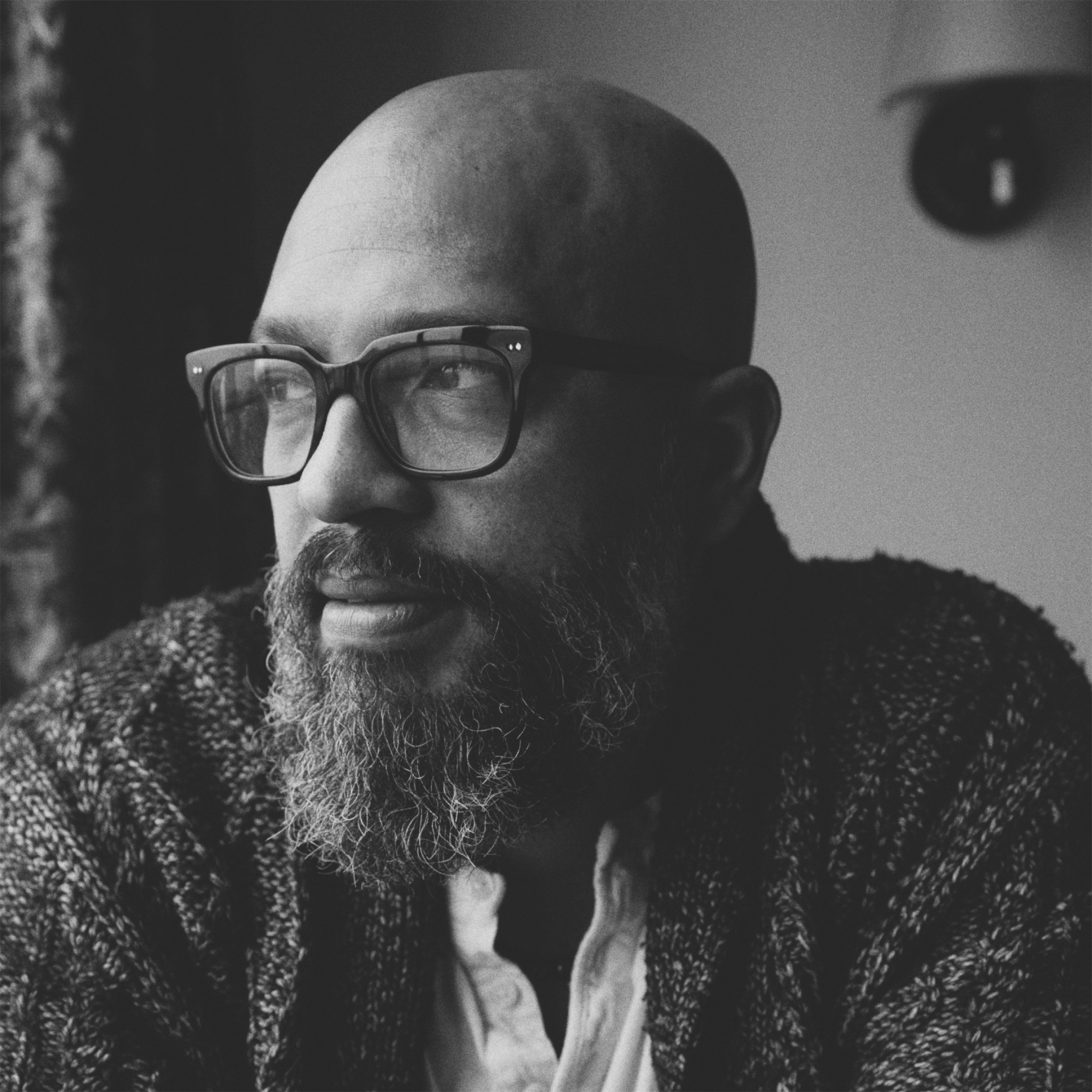 A man with dark, neatly styled hair and a trimmed beard wears rectangular glasses and a tweed jacket over a collared shirt. He stands in front of a plain wall and looks directly at the camera with a slight smile. The image is in black and white.