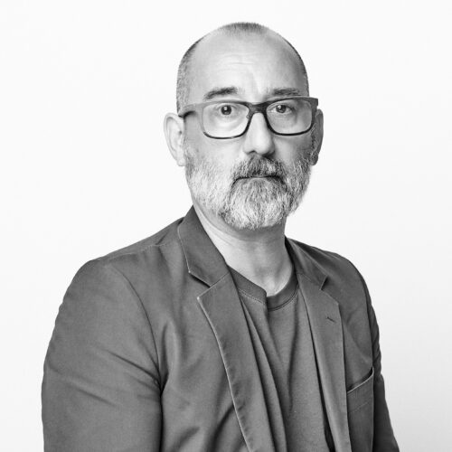 Black-and-white portrait of a man with a close-cropped haircut, beard, and glasses, wearing a blazer over a T-shirt. He sits facing the camera with a neutral expression against a plain white background.