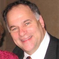Portrait of a smiling man in a suit and tie, captured indoors against a softly lit brown background. He has short dark hair graying at the sides and is shown from the shoulders up, leaning slightly toward the camera.