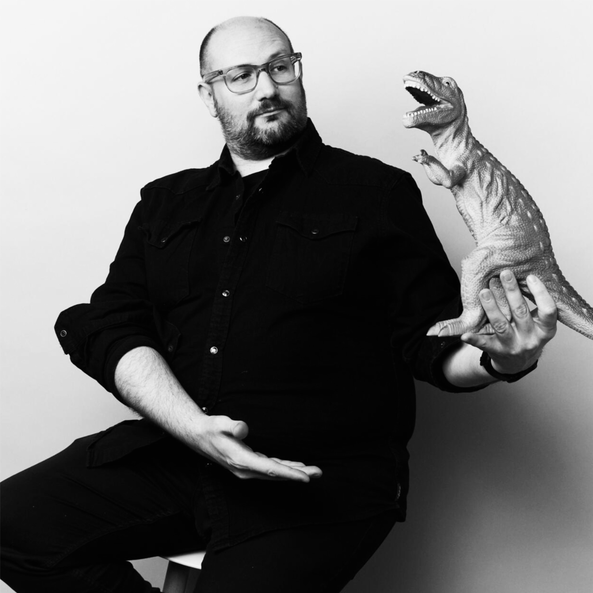 A man with glasses and a beard, dressed in a dark button-up shirt, sits on a stool and looks toward a toy dinosaur perched on his hand. The image is black and white with a playful tone, suggesting a mix of seriousness and humor.