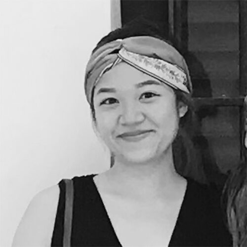 A smiling woman wearing a patterned headband and sleeveless black top poses indoors. She stands in front of a white wall and a wooden shelf. Her expression is warm and friendly. The photo is in black and white.
