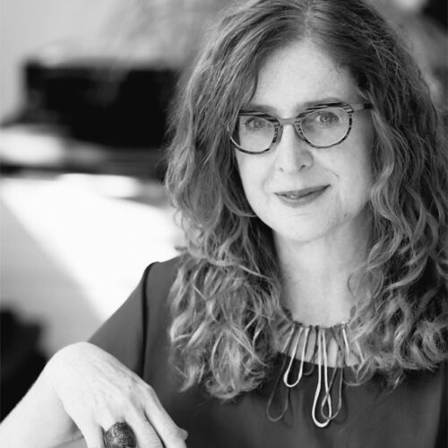 A woman with long curly hair and glasses sits indoors, smiling softly at the camera. She wears a dark blouse and a statement necklace made of large metal loops. The background includes a piano and blurred indoor elements. The photo is in black and white.