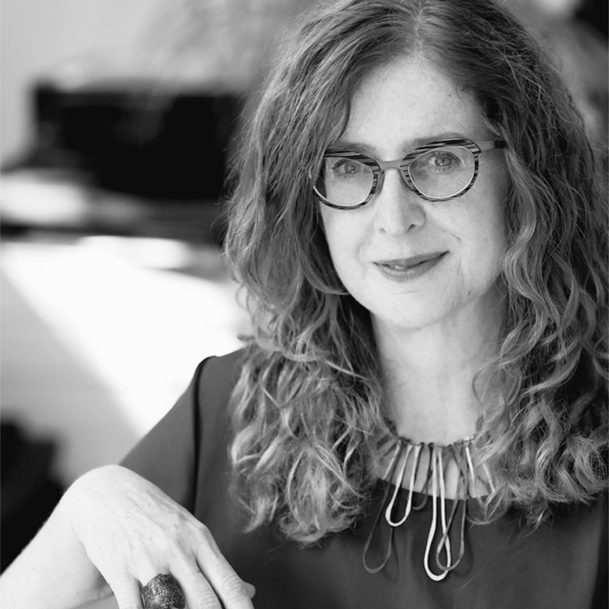 A woman with long curly hair and glasses sits indoors, smiling softly at the camera. She wears a dark blouse and a statement necklace made of large metal loops. The background includes a piano and blurred indoor elements. The photo is in black and white.