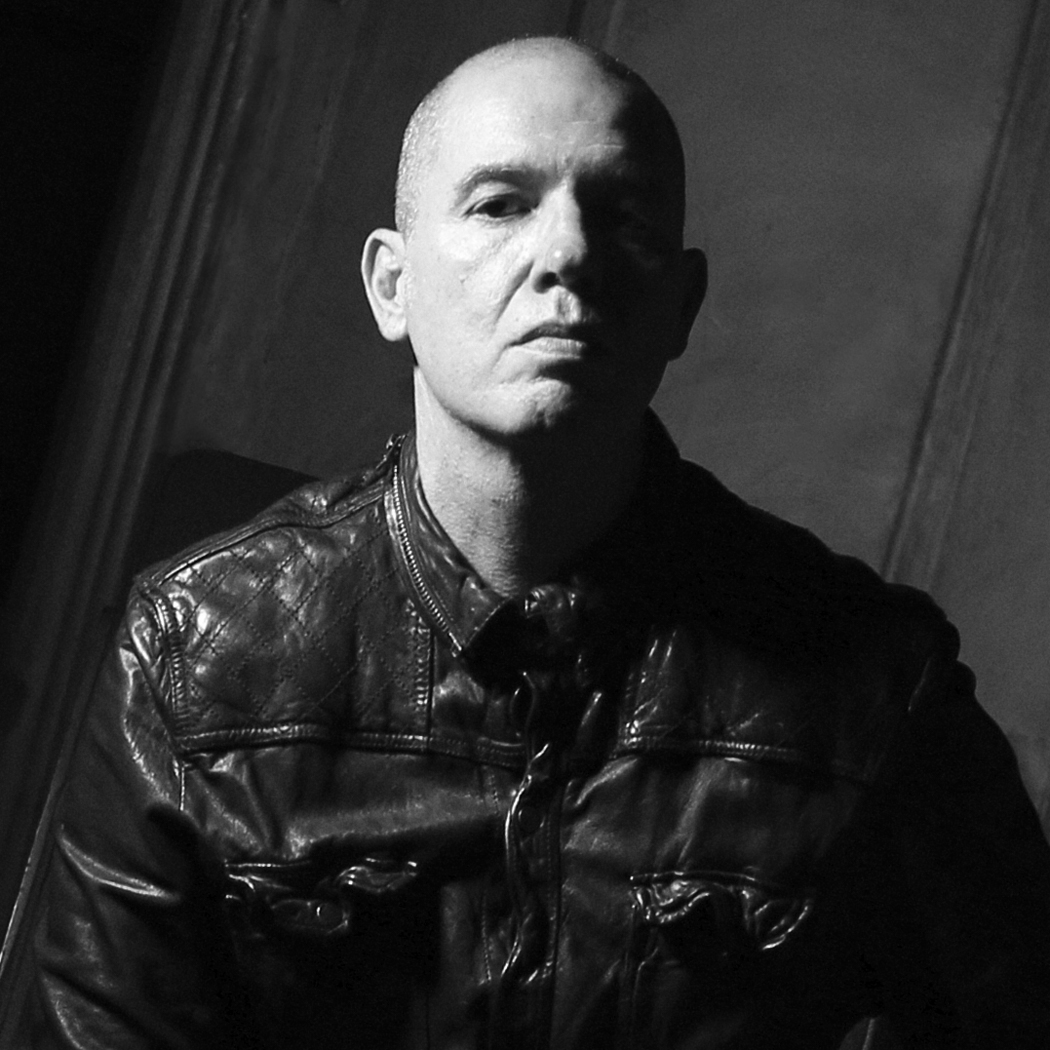 A bald man in a quilted leather jacket stares forward with a serious, almost stoic expression. He is seated in a dimly lit interior with shadows enhancing the textured wall behind him. The image is shot in high-contrast black and white.