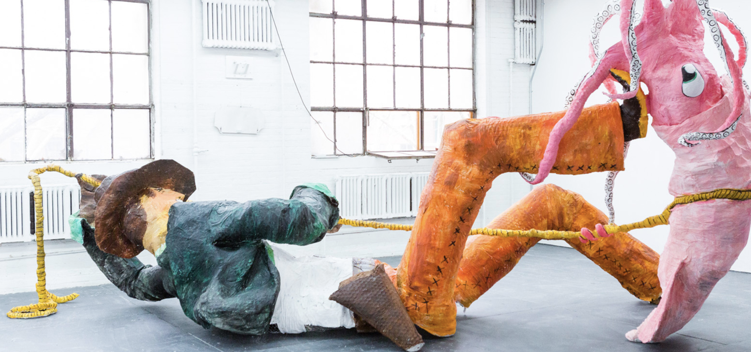 A sculpture of a cowboy on his back lassoing a large pink kraken installed in a white room with large windows.