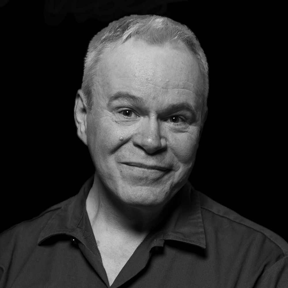 A man with short, silver hair smiles gently at the camera. He wears a collared shirt and is lit from the front against a black background. The black and white portrait emphasizes warmth and approachability.