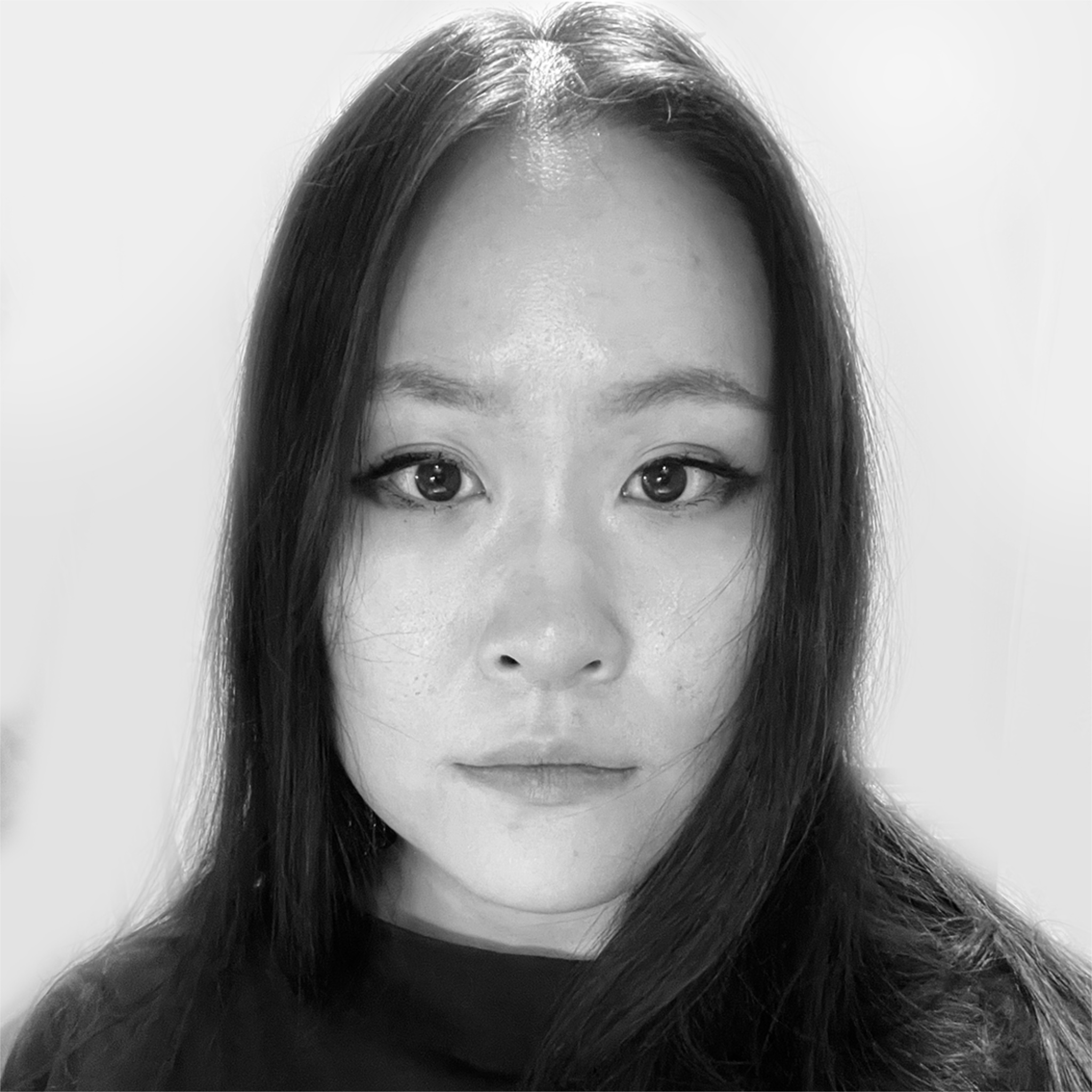A woman with long black hair stares directly at the camera with a neutral expression. She wears a dark top, and her surroundings are softly lit with a light gray background. The black and white photo emphasizes facial features and intensity.