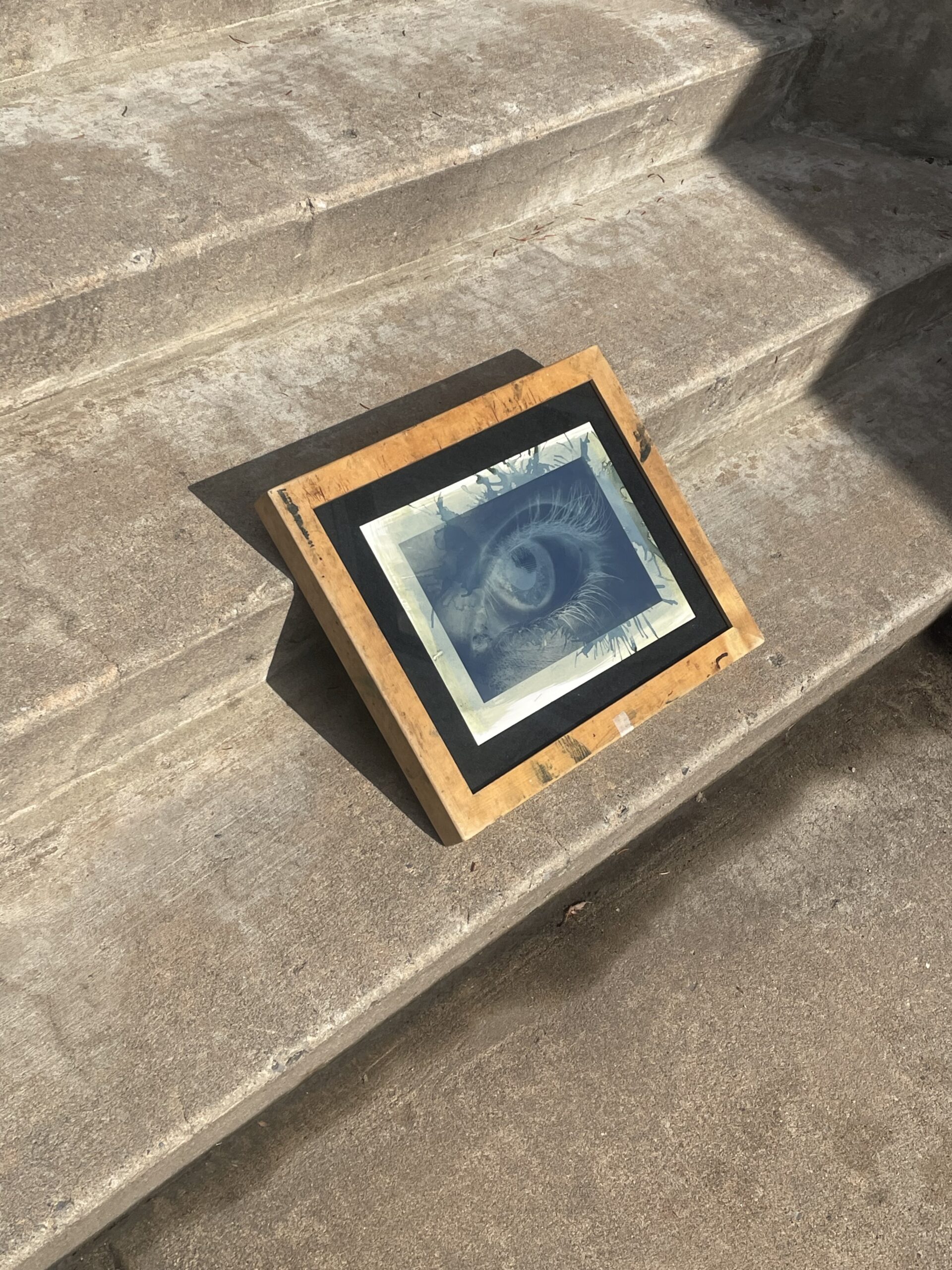 A framed artwork featuring an eye is placed on a set of stone steps. The frame is made of a light-colored wood with a black accent around the artwork. The steps appear weathered and are illuminated by sunlight.