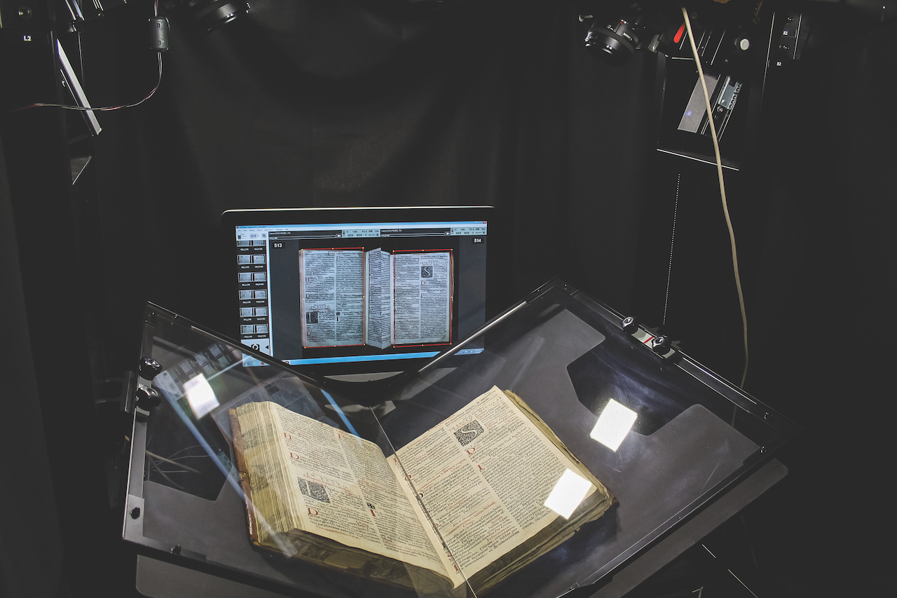 photo scanning and archiving of a large book, done with multiple cameras, a video monitor and lighting setup, book is behind thick protective glass