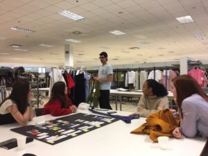 professor speaking to students, in a clothes workshop, describing clothes on hangers to other students, other clothes on hangers in the background