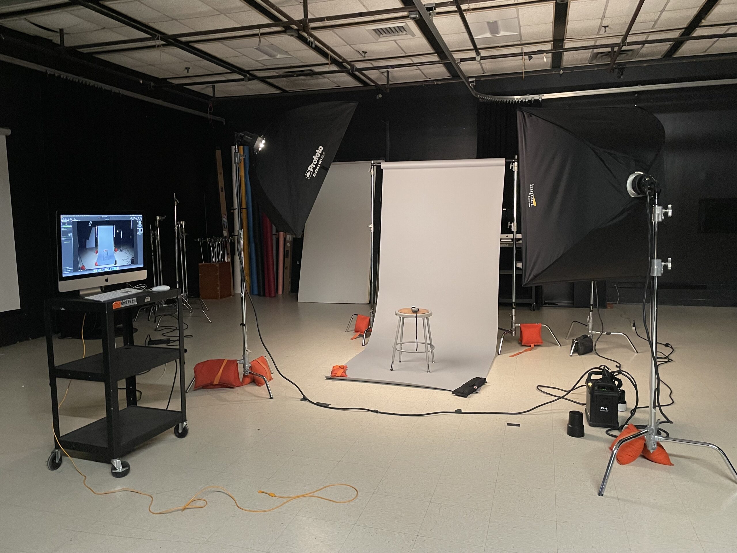 A photography studio setup featuring softbox lights on either side, a gray backdrop, and a stool positioned in front. A computer monitor on a cart displays the live feed from the camera. The floor is covered with light-colored tiles, and various lighting equipment is arranged around the space, with cables visible on the ground.