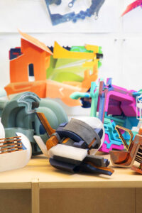 Colorful, abstract architectural models arranged on a tabletop, featuring layered geometric forms in wood, plastic, and painted materials. The foreground shows sculptural components in blue, white, gray, and natural wood tones, while the background includes brightly colored cut-paper or foam-core studies pinned to the wall. The scene conveys an active design studio environment with experimental material exploration.
