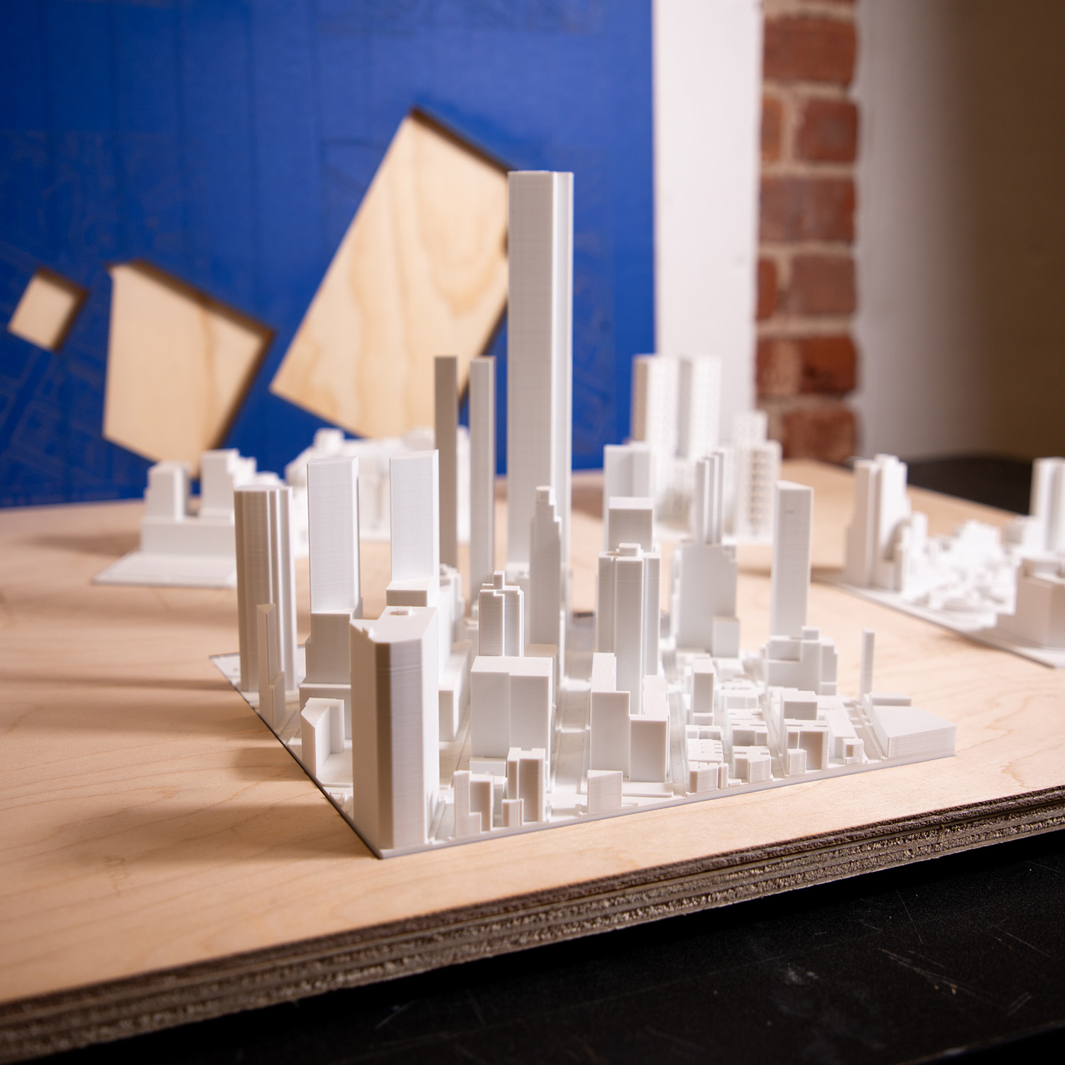 Photograph of a white 3D-printed architectural city model displayed on a wooden table. The model features numerous simplified building forms of varying heights, including several tall tower structures rising above smaller surrounding blocks. In the background, additional models and geometric wood panels are visible against a blue wall and exposed brick, suggesting a design studio or academic setting.