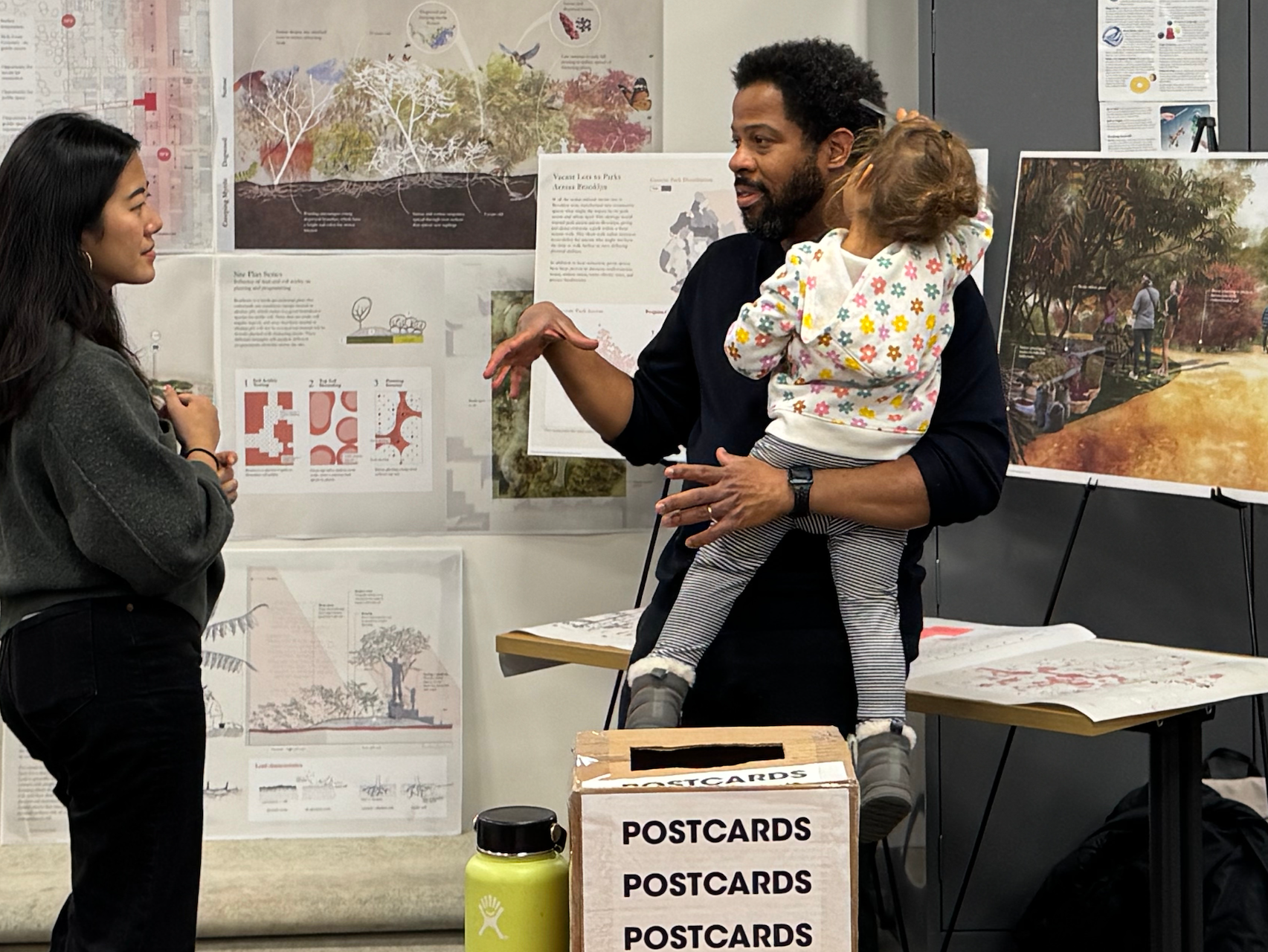 Indoor exhibition or studio setting with three people engaged in conversation. One adult stands at right holding a small child on their hip while gesturing mid-discussion; the child wears patterned clothing and looks away from the camera. Another adult stands at left listening attentively. Behind them, multiple architectural or landscape design posters and drawings are pinned or mounted on the wall, with a large illustrated rendering on an easel to the right. In the foreground, a cardboard box labeled “POSTCARDS” sits on a table alongside a reusable water bottle, indicating a public presentation, critique, or open studio environment.