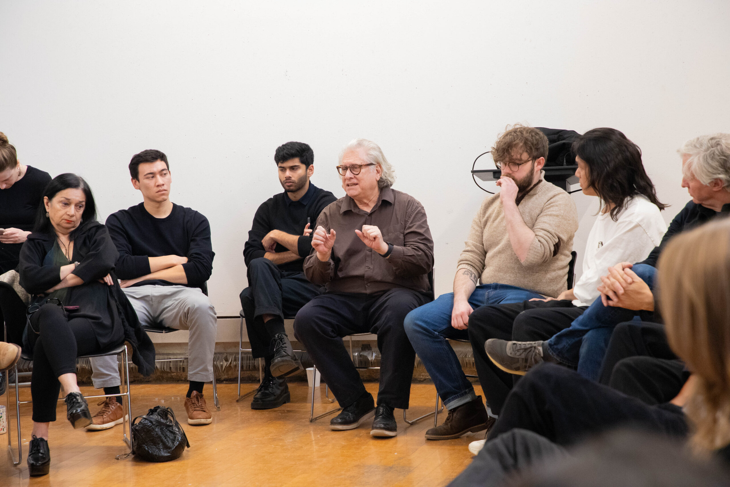 A group of adults sit in a circle during a discussion or critique session. One older man in the center gestures with his hands while speaking. Others sit attentively around him in chairs within a studio space with plain walls and wooden flooring.