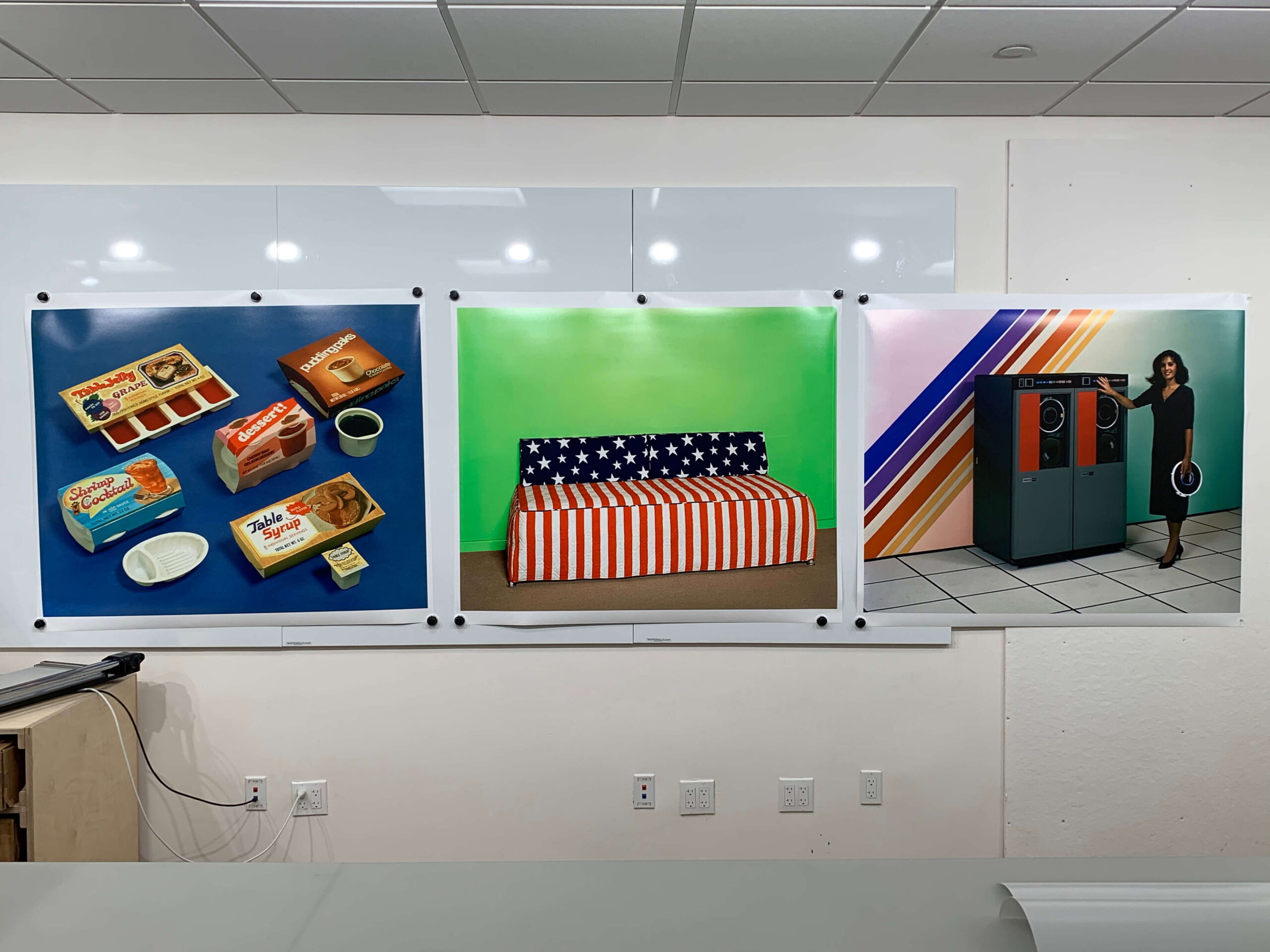 A white wall has three photos mounted on it. The first photo is of several food products with colorful packaging against a blue background. The second is of a bed with American flag bedsheets on it. There is a green screen behind the bed. The third picture is of a saleswoman presenting a large electronic device.
