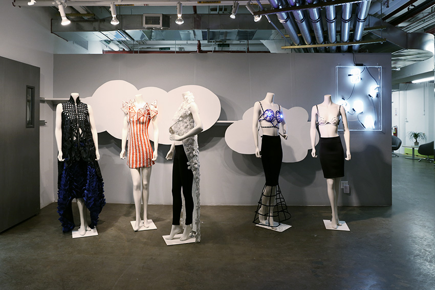 Five mannequins stand in front of a grey wall. They are each wearing different garments and facing in aleatory directions.