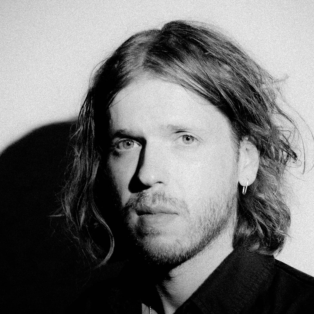 A black and white portrait of a man with shoulder-length wavy hair and facial hair. He wears a button-up shirt and a hoop earring, gazing seriously at the camera. The lighting casts dramatic shadows on the background wall.