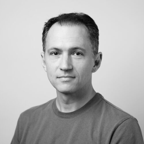A grayscale headshot of a man with short, dark hair, wearing a plain crew neck shirt. He looks directly at the camera with a composed and neutral expression against a plain, light background.