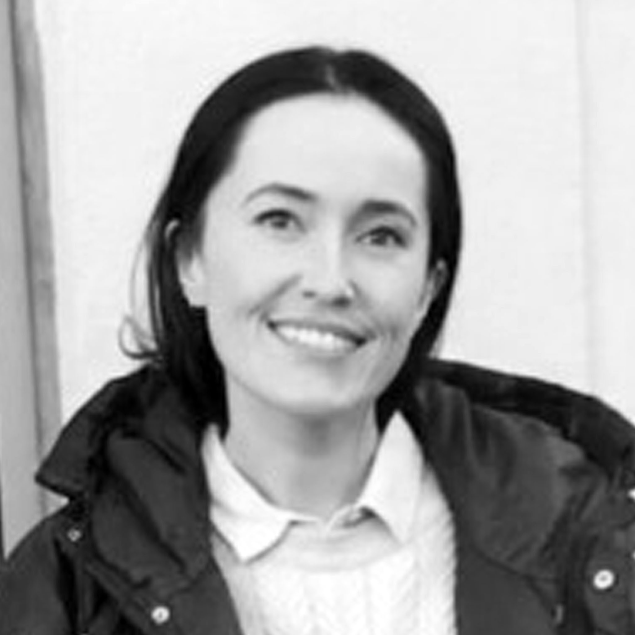 A woman stands smiling in front of a pale wall, wearing a dark coat over a light cable-knit sweater and collared shirt. Her dark hair is shoulder-length and tucked behind her ears. The image is bright and friendly.