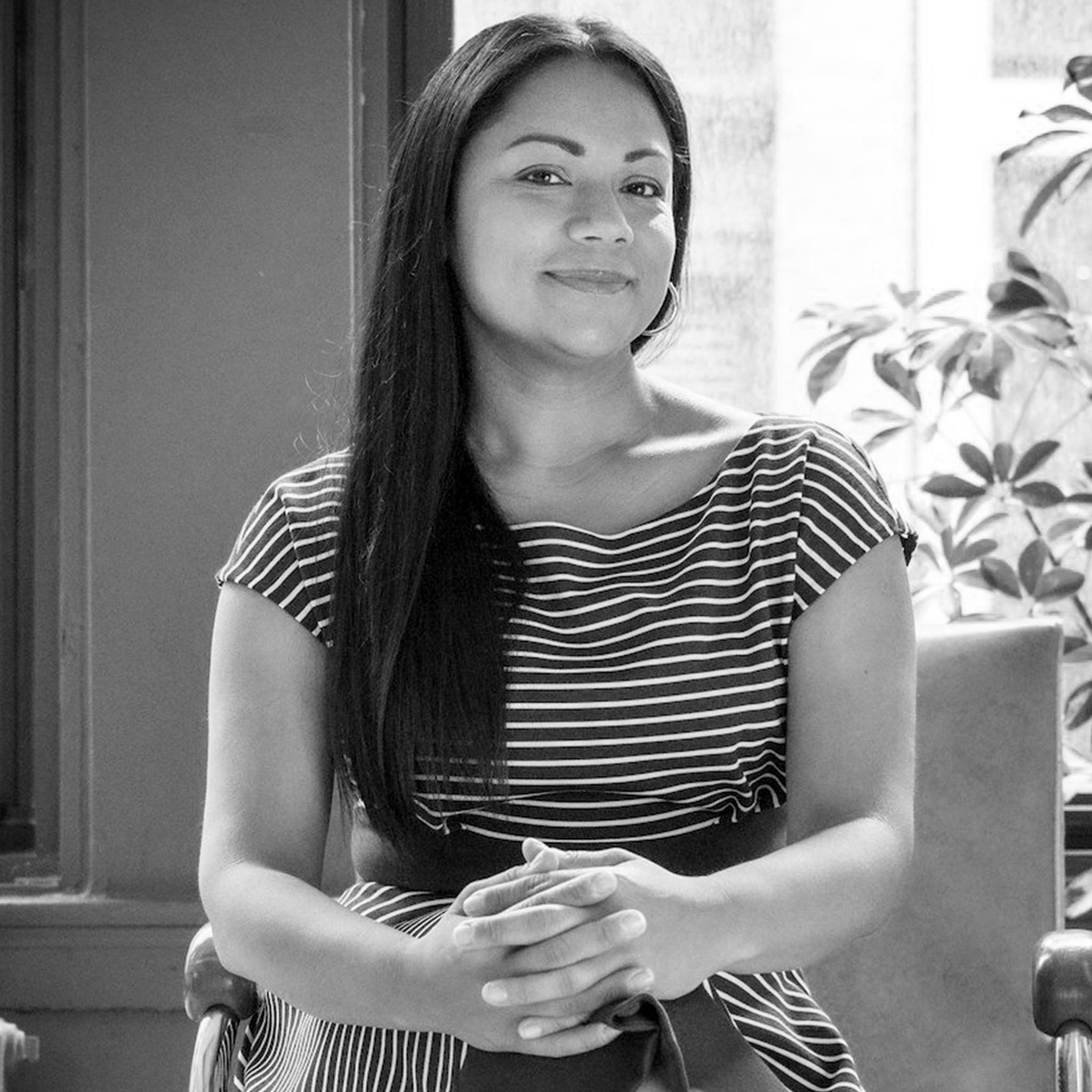 A woman with long straight hair sits indoors, hands folded in her lap. She wears a striped dress with cap sleeves and smiles softly. Behind her is a window and a potted plant. The black and white photo feels composed and professional.