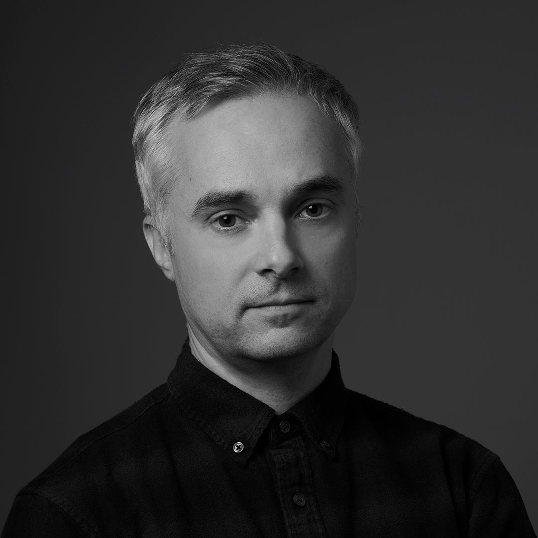A man with neatly combed gray hair and a calm expression wears a dark button-up shirt. He is facing slightly to the side against a dark background. The portrait is in black and white with soft lighting.