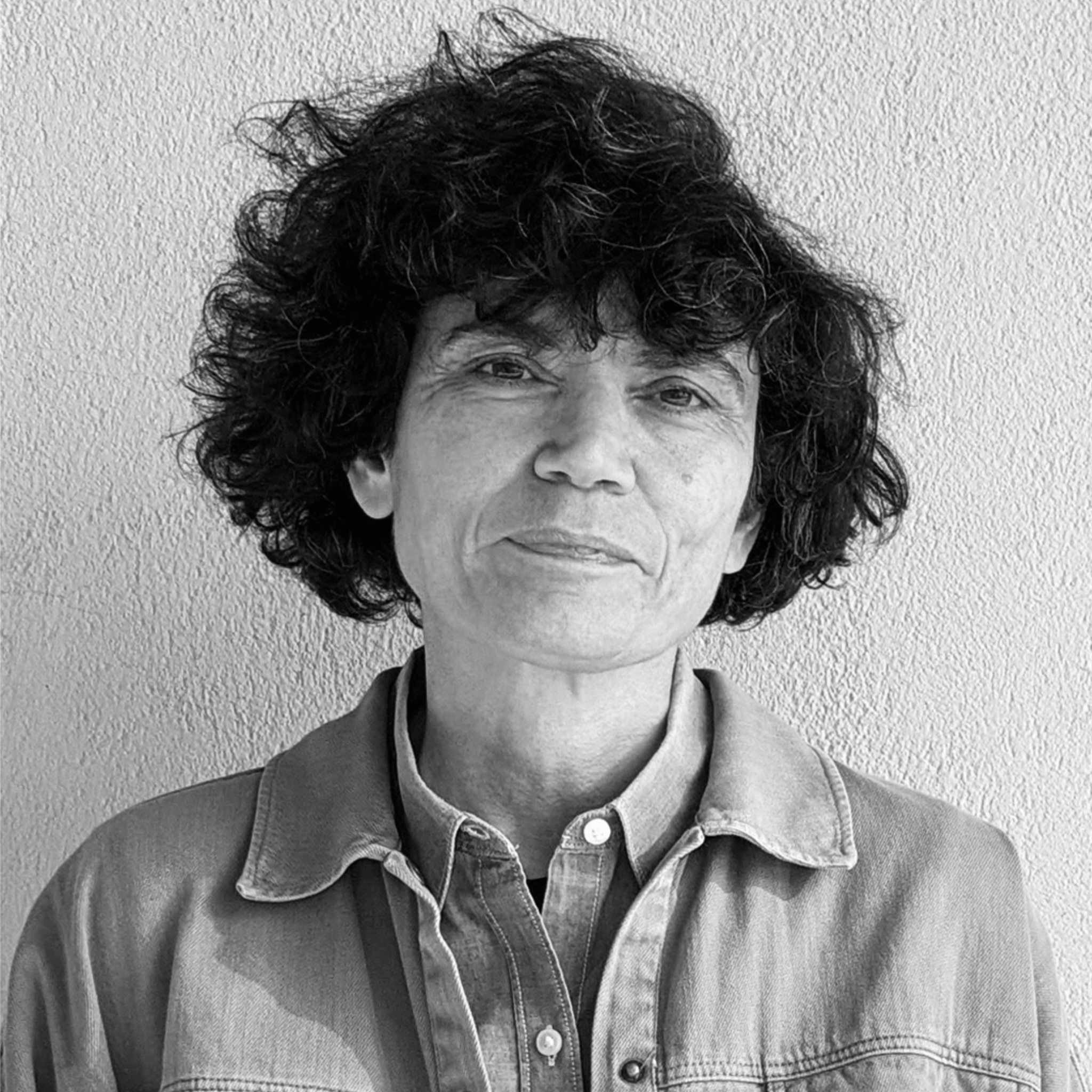 A woman with curly dark hair and a calm expression stands in front of a textured light-colored wall. She wears a buttoned shirt layered under a denim jacket. The photograph is in black and white with soft natural lighting.