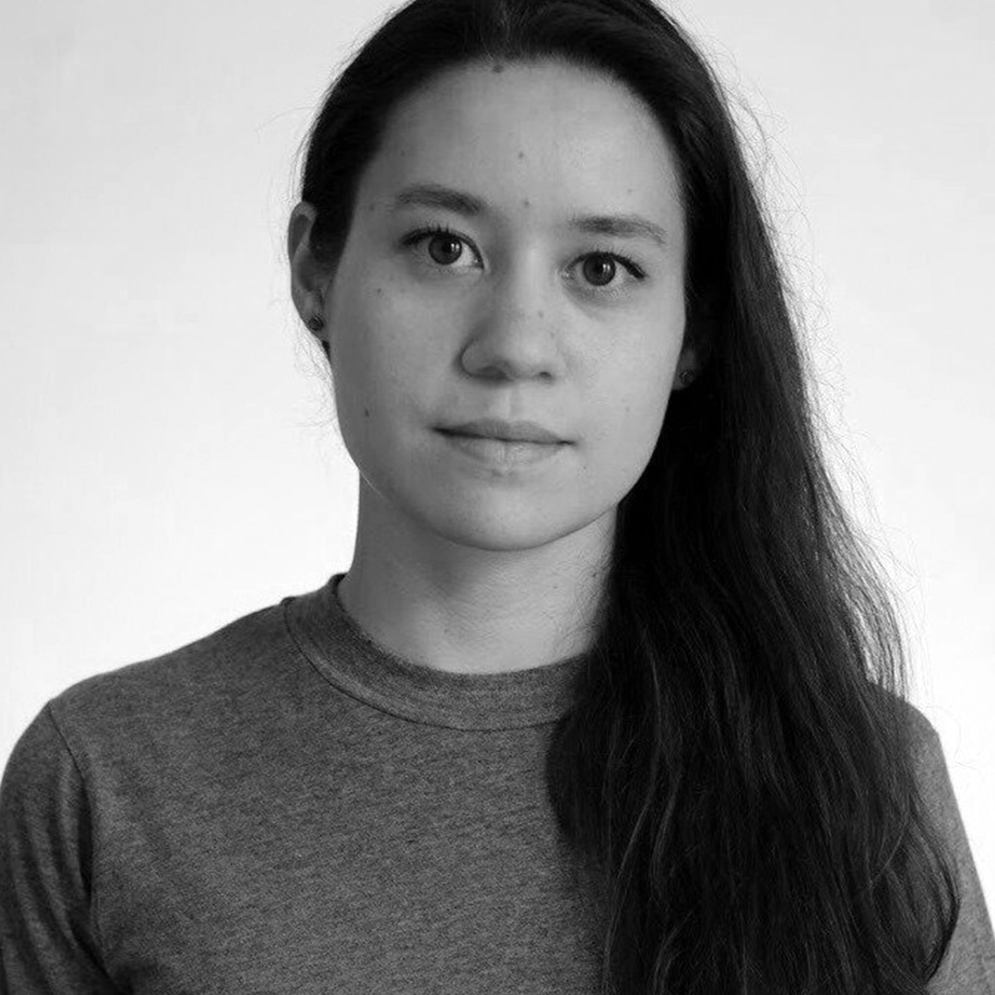 A woman with long dark hair stands against a plain white background. She wears a simple t-shirt and gazes directly at the camera with a neutral, composed expression. The image is straightforward and minimal.