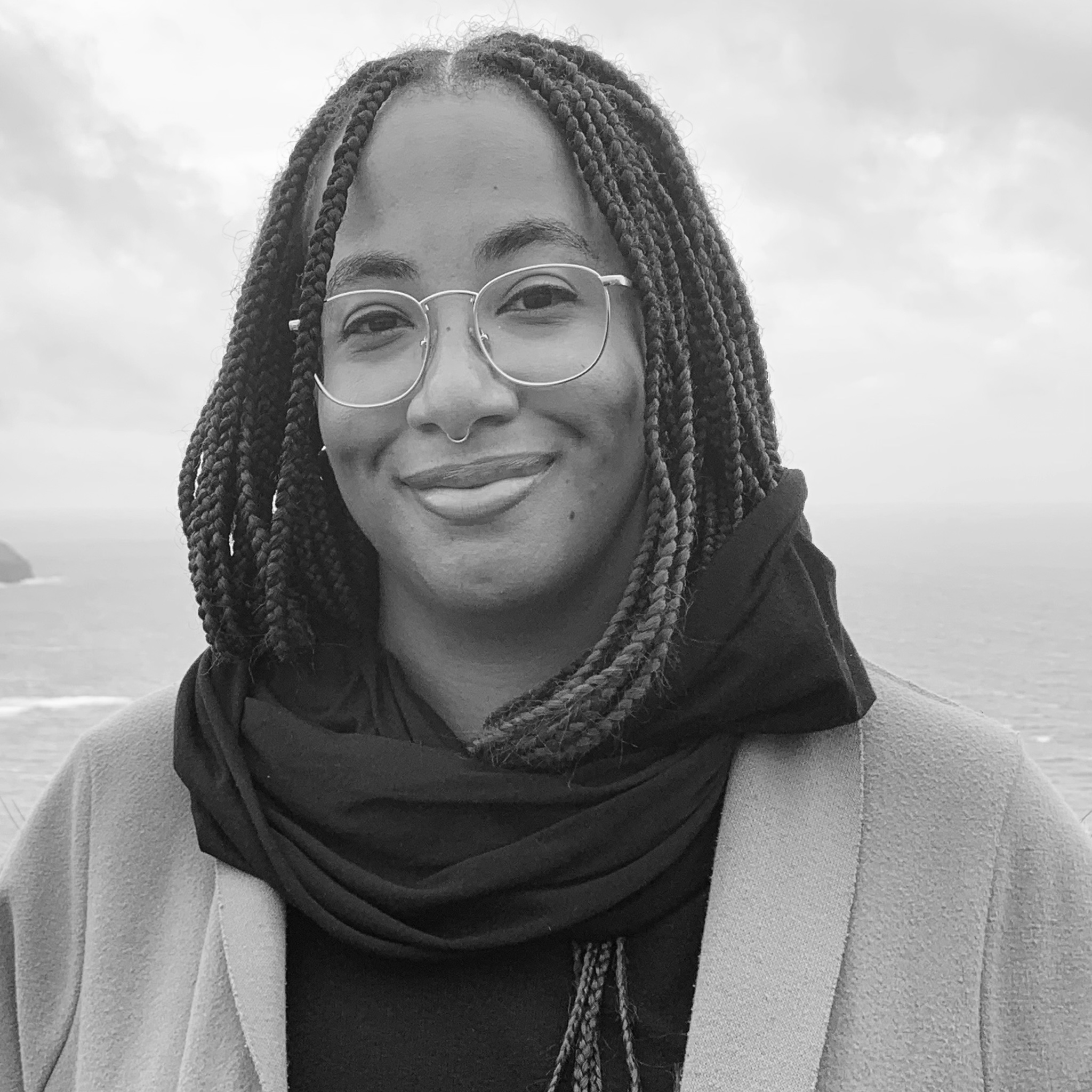 A woman with long braided hair and round glasses smiles at the camera. She wears a scarf and jacket, standing outdoors near a coastal cliff with the ocean and cloudy sky behind her. The image is black and white and serene.