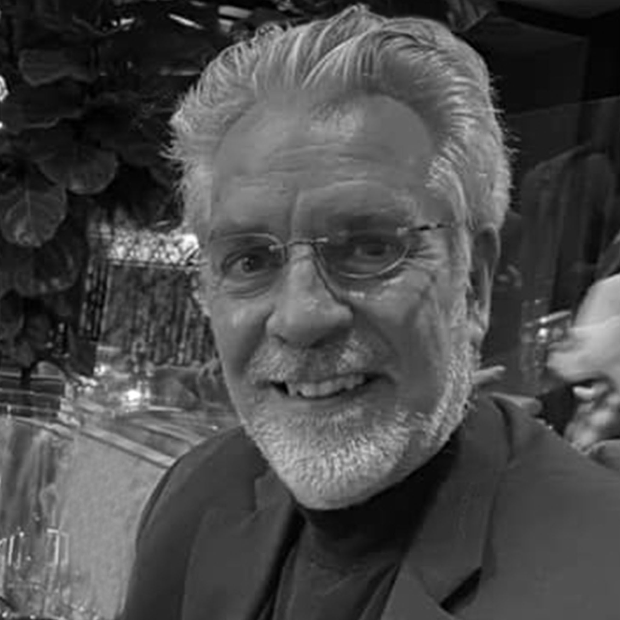 A man with short curly hair and a beard sits in a bright indoor space, wearing a plaid button-up shirt. He smiles with a relaxed demeanor, and the photo’s high contrast and lighting emphasize a casual, professional presence.