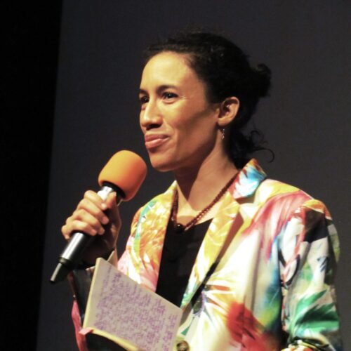 Layla Zami gives a speech holding a microphone in one hand and a notebook in another. She is wearing a colorful blazer with parrots printed on it