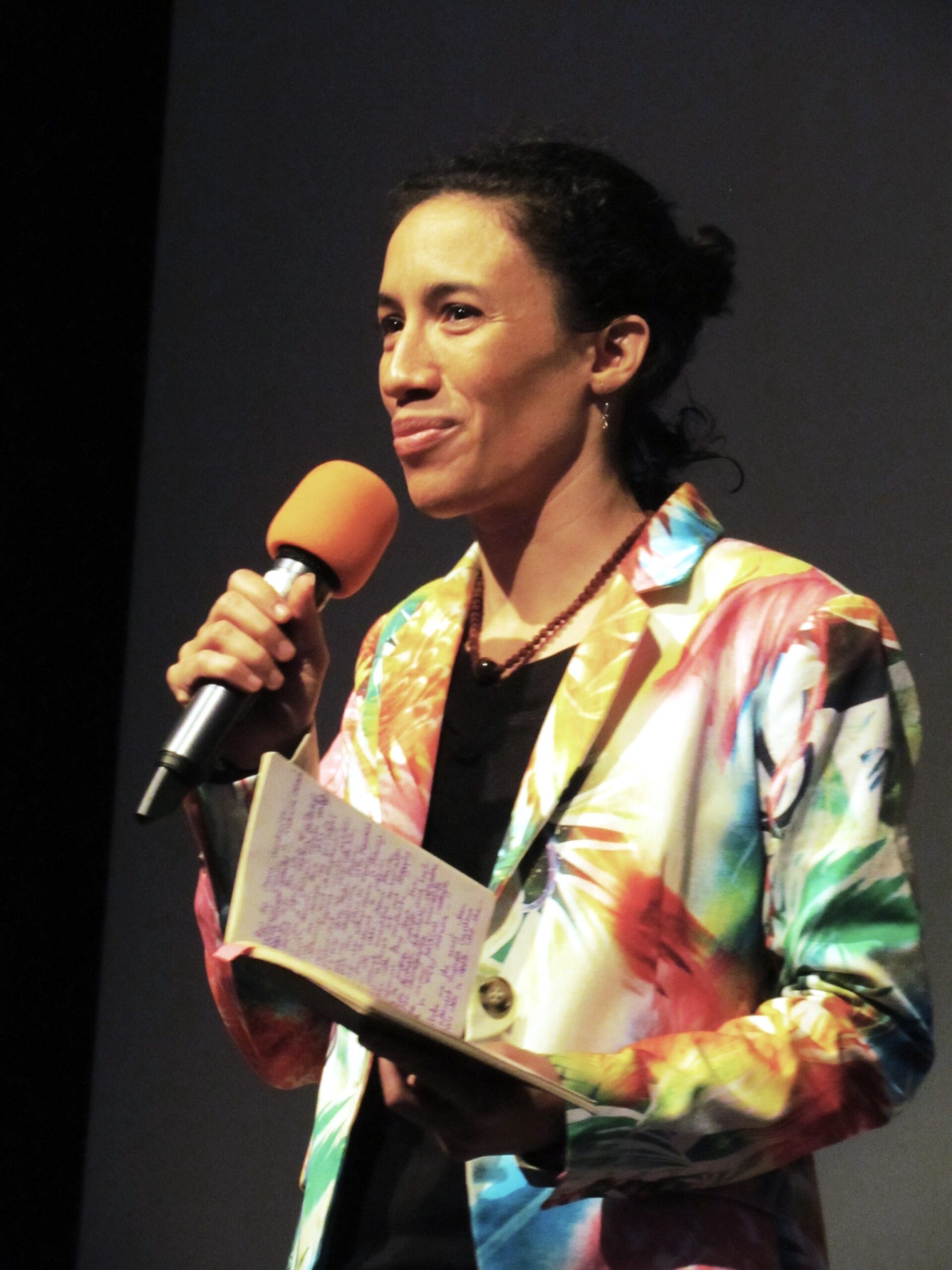 Layla Zami gives a speech holding a microphone in one hand and a notebook in another. She is wearing a colorful blazer with parrots printed on it
