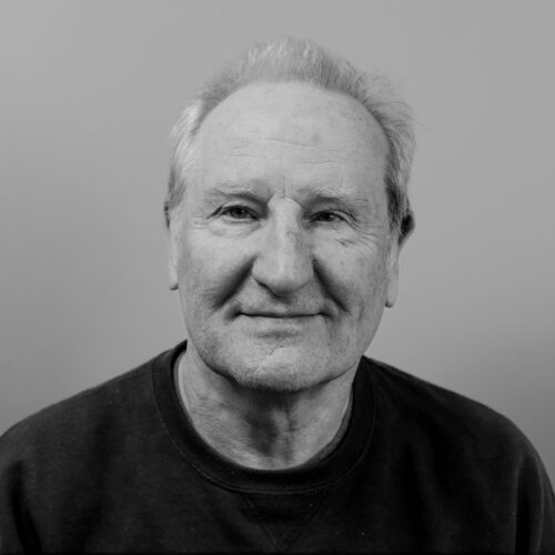 Black-and-white portrait of an older man with short, light-colored hair, wearing a dark crewneck shirt. He faces the camera directly with a calm, faint smile, against a plain, softly lit background that emphasizes his facial expression and texture.