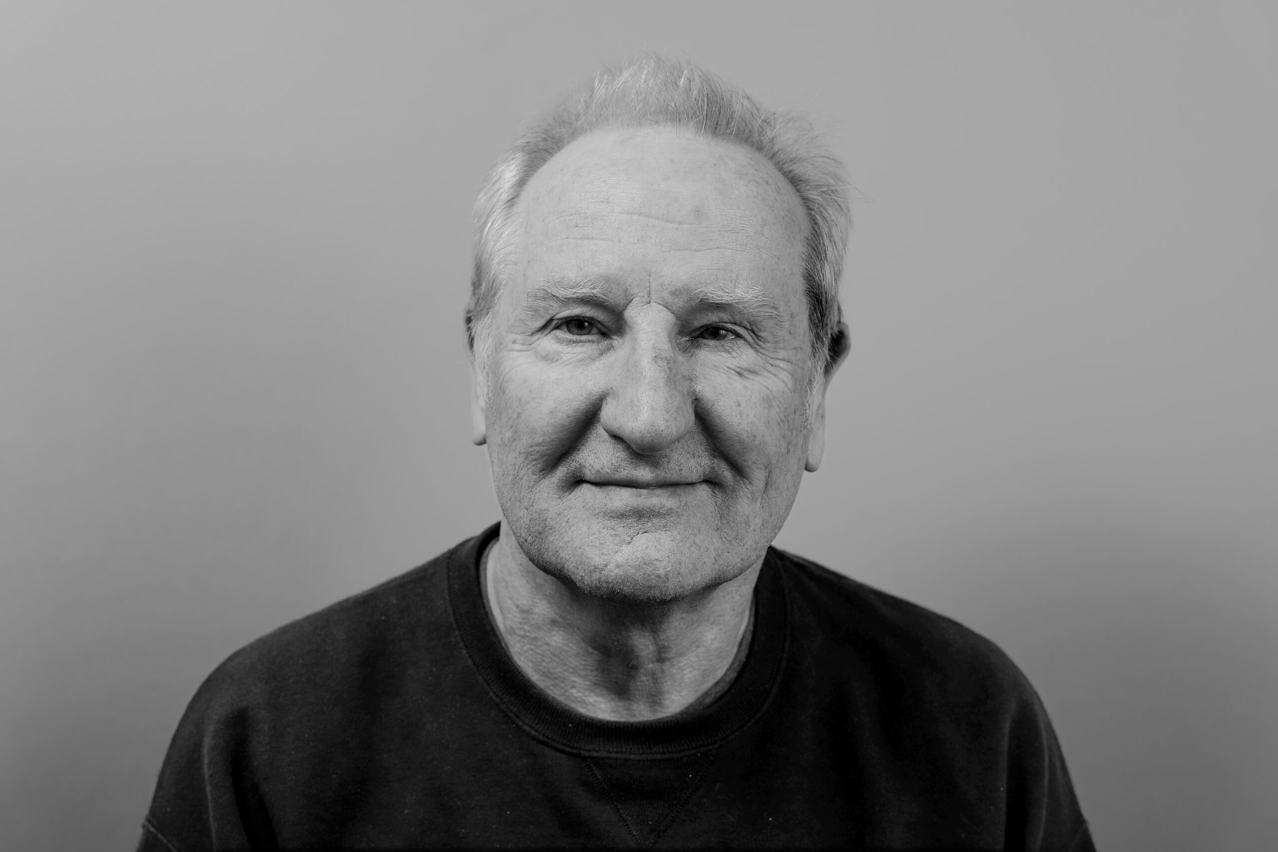 Black-and-white portrait of an older man with short, light-colored hair, wearing a dark crewneck shirt. He faces the camera directly with a calm, faint smile, against a plain, softly lit background that emphasizes his facial expression and texture.