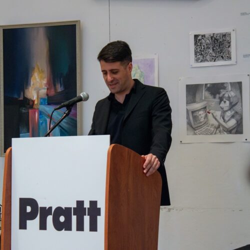A man in a black jacket stands at a podium with the word "Pratt" prominently displayed. He is speaking into a microphone, appearing to be engaged and smiling. Behind him, artwork hangs on the wall, including colorful abstract pieces and detailed black-and-white drawings.
