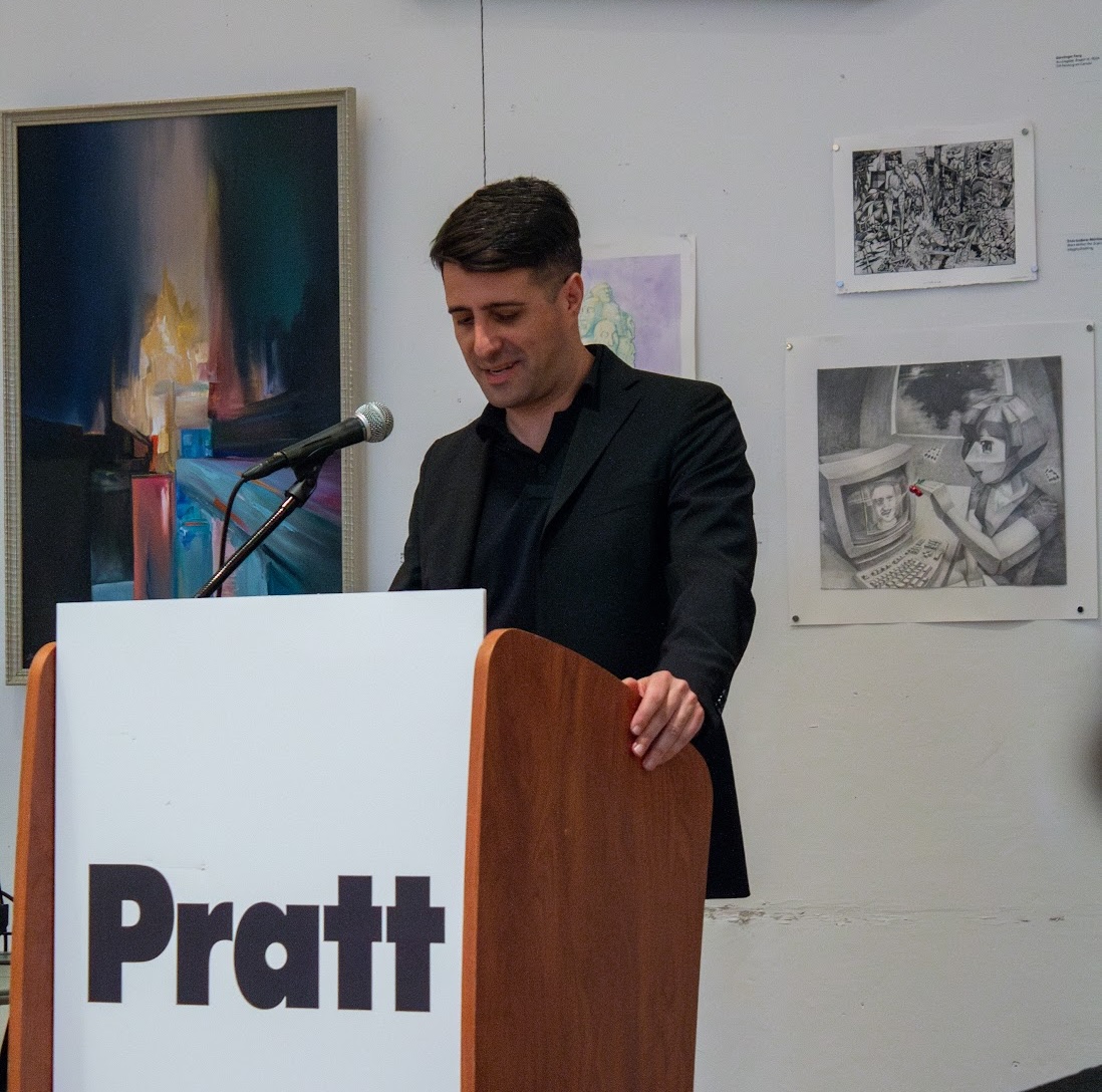 A man in a black jacket stands at a podium with the word "Pratt" prominently displayed. He is speaking into a microphone, appearing to be engaged and smiling. Behind him, artwork hangs on the wall, including colorful abstract pieces and detailed black-and-white drawings.