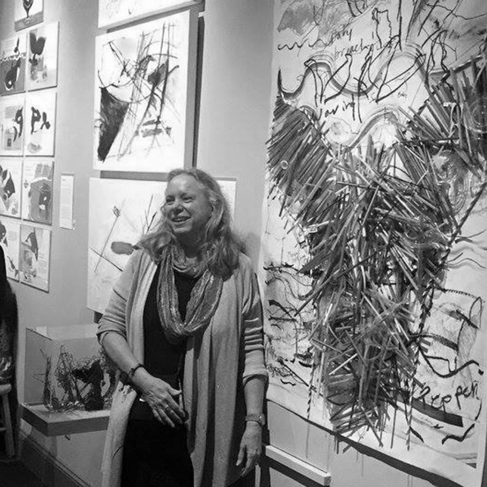 A woman stands in front of a gallery wall covered with expressive, abstract artwork. She has long, light-colored hair and wears a scarf and cardigan. She smiles as she engages with someone or something off-frame.