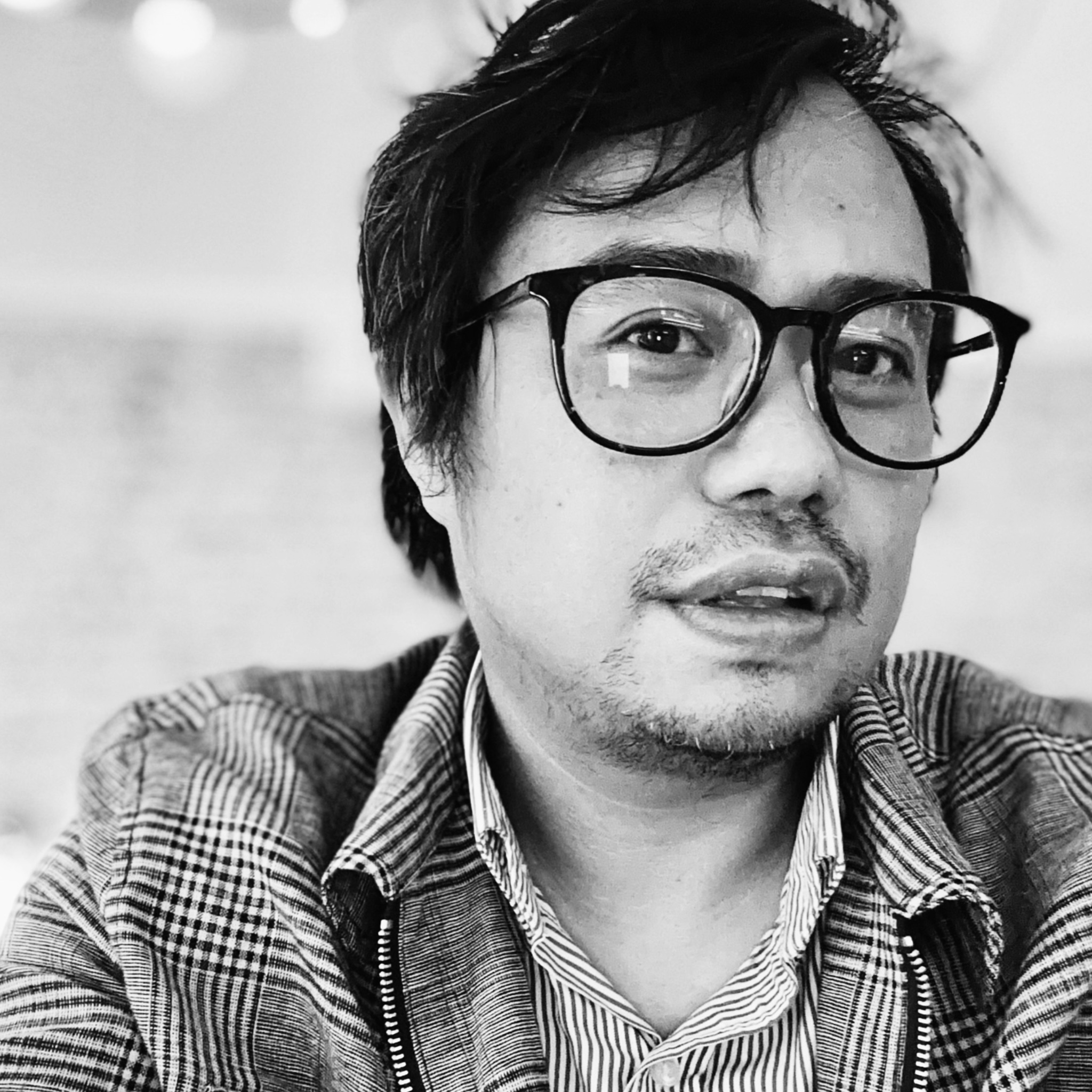 A man with tousled dark hair, a short beard, and rectangular glasses gazes past the camera. He wears a patterned jacket over a striped shirt. The background is softly blurred, with string lights and indoor décor faintly visible. The image is black and white.