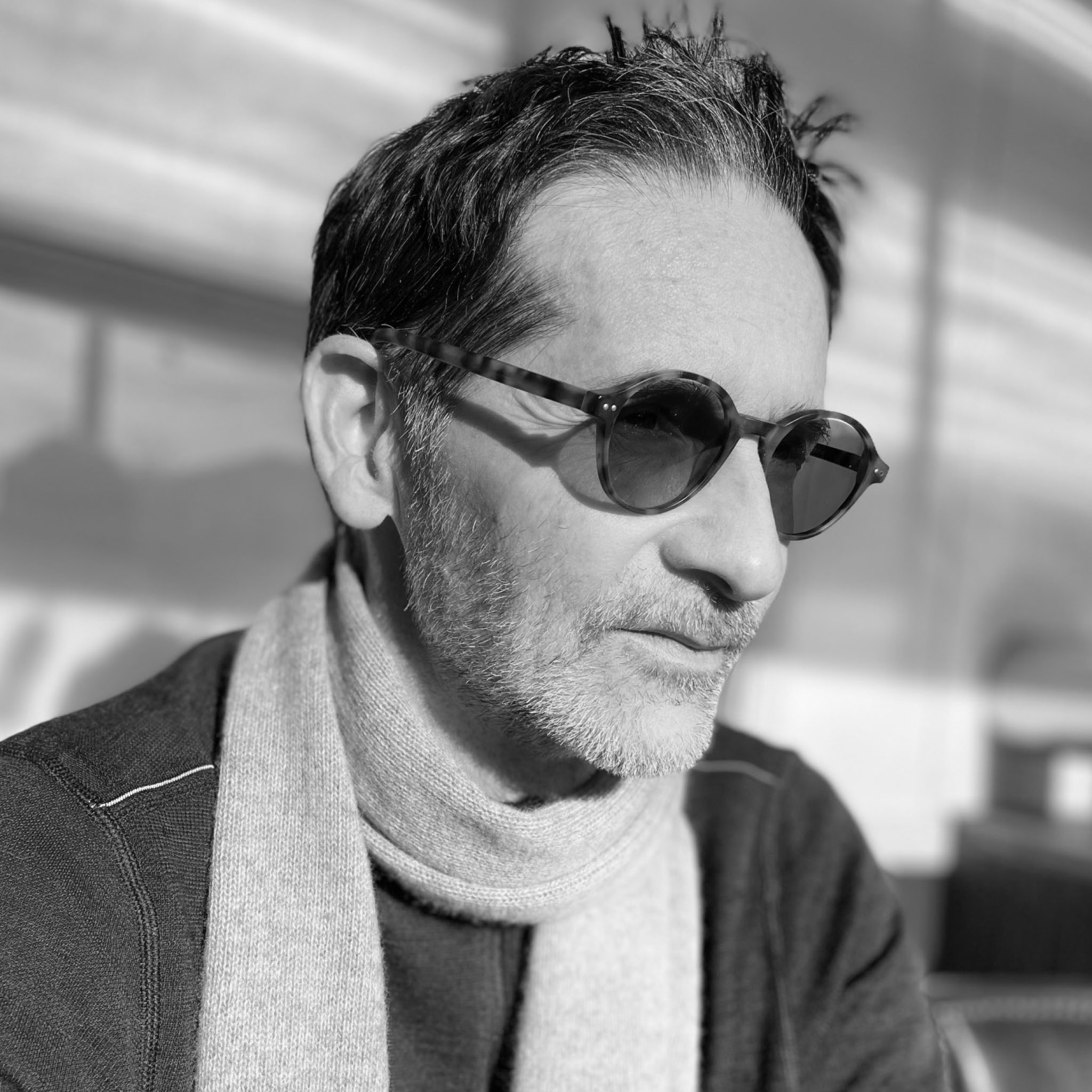 A man with short dark hair and a salt-and-pepper beard looks to the side while wearing sunglasses and a scarf. The photo is softly lit, giving a casual and reflective tone to this black and white portrait.