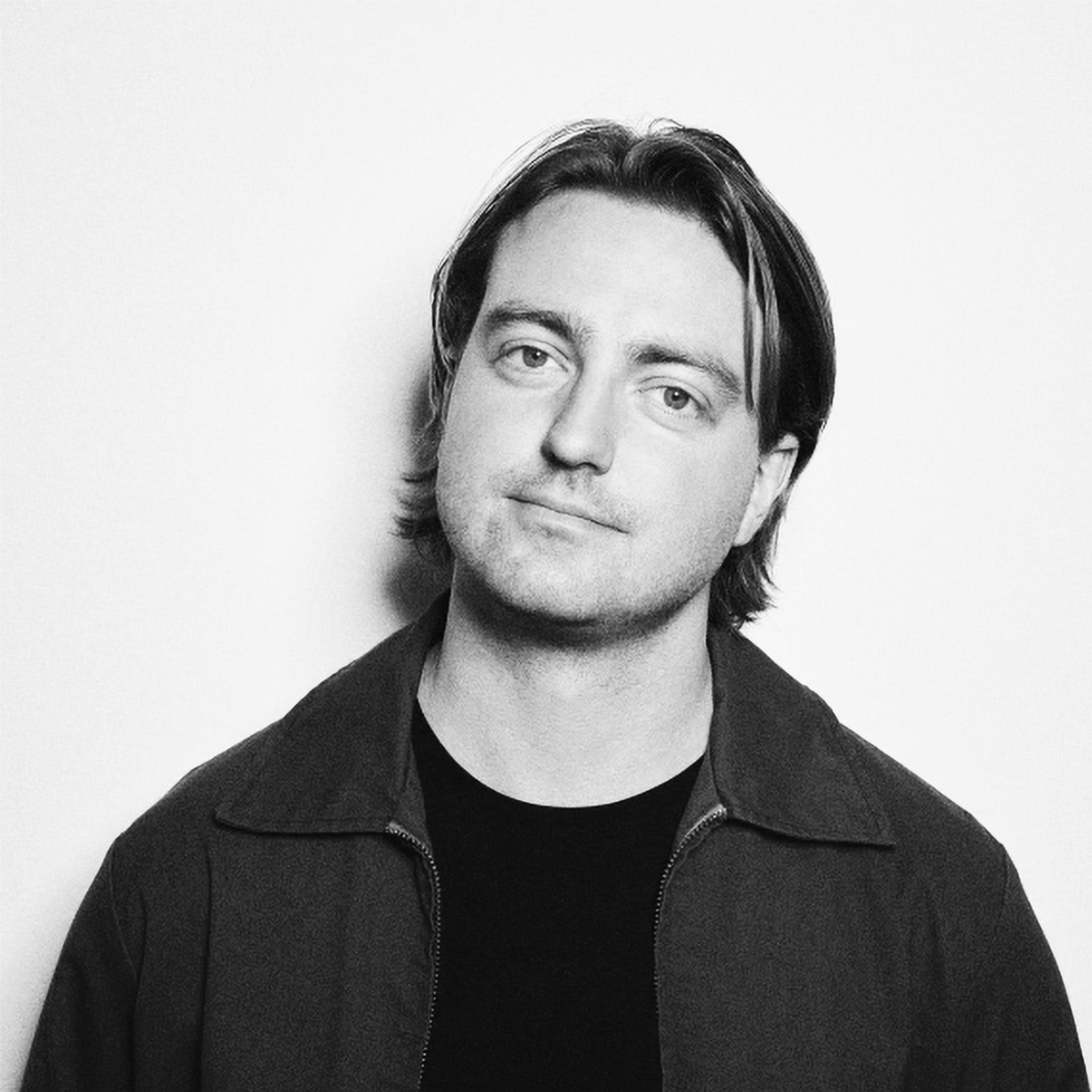 A man with shoulder-length hair and a slight smile stands against a white background. He wears a dark crew-neck shirt under a jacket. His relaxed posture and direct gaze give a casual, approachable impression. The image is in black and white.